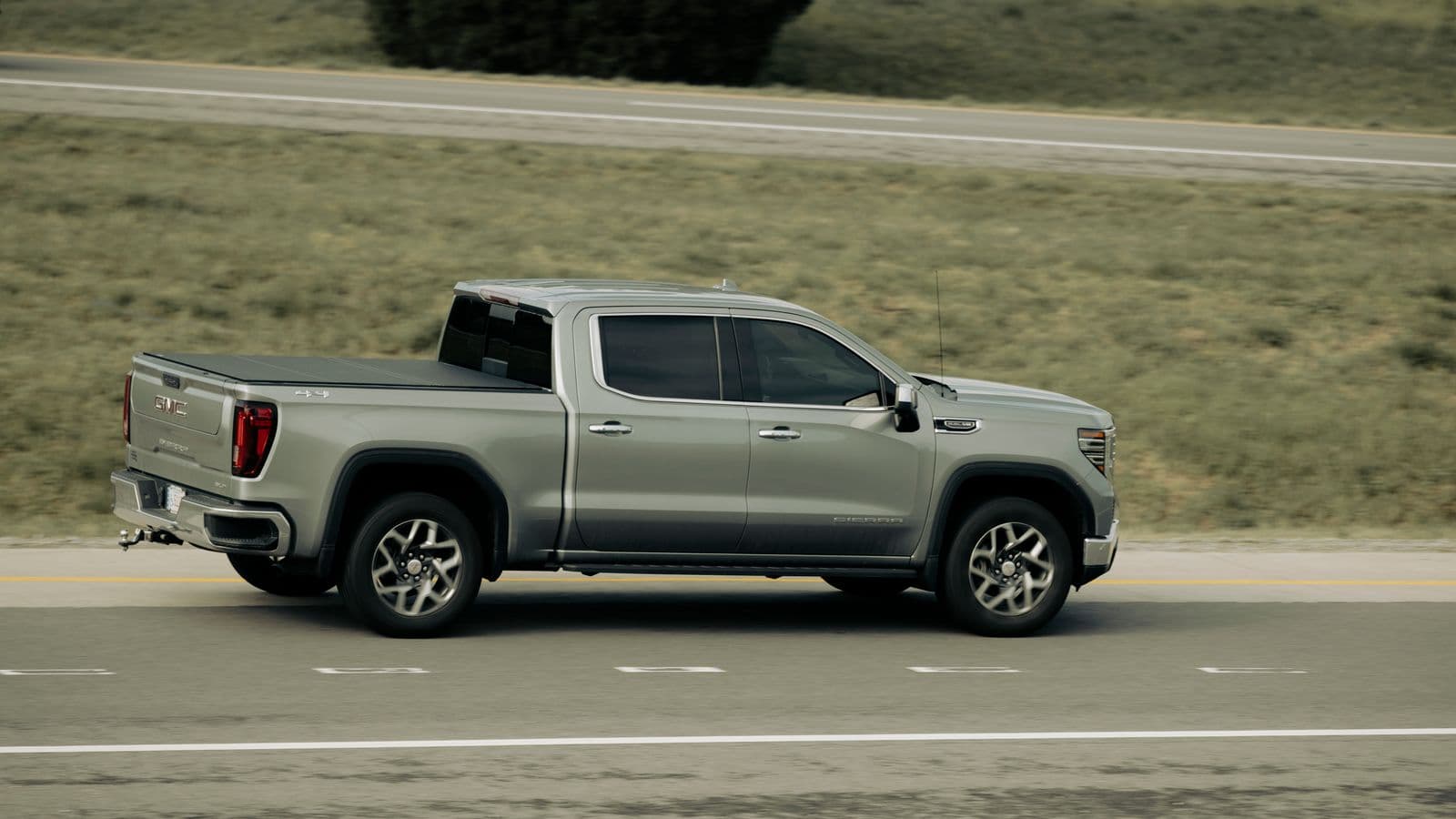 A silver GMC Sierra 1500 crew cab on the highway with a flat low-profile hard tonneau cover installed